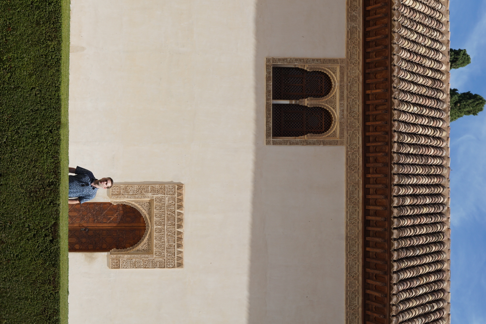 Strong diagonal shadows across the white plasterwork of the Alhambra, a figure passing beneath an ornate arch.