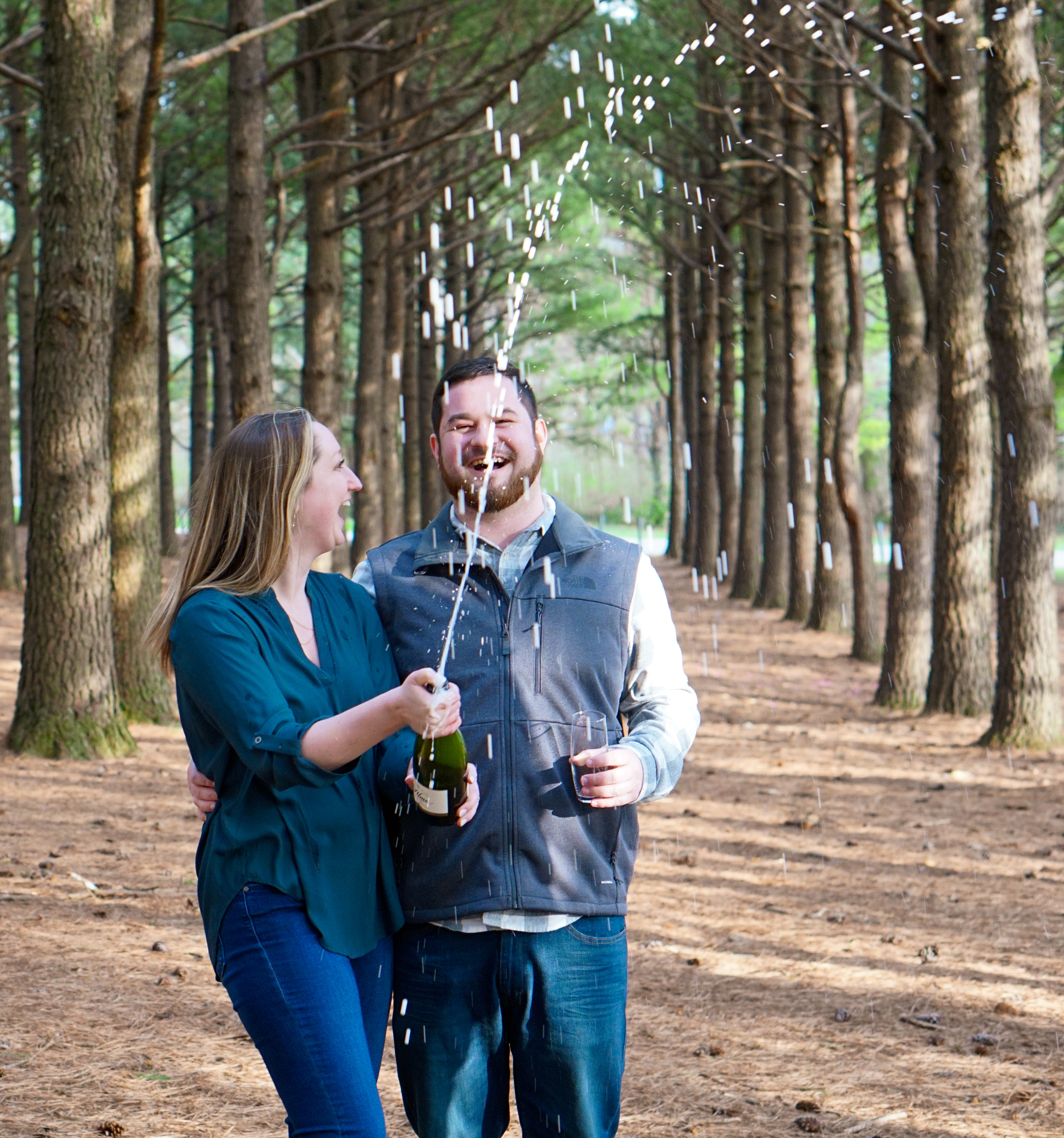 Couple laughing and popping champagne in a sunlit pine tree avenue during an engagement shoot in Lexington KY