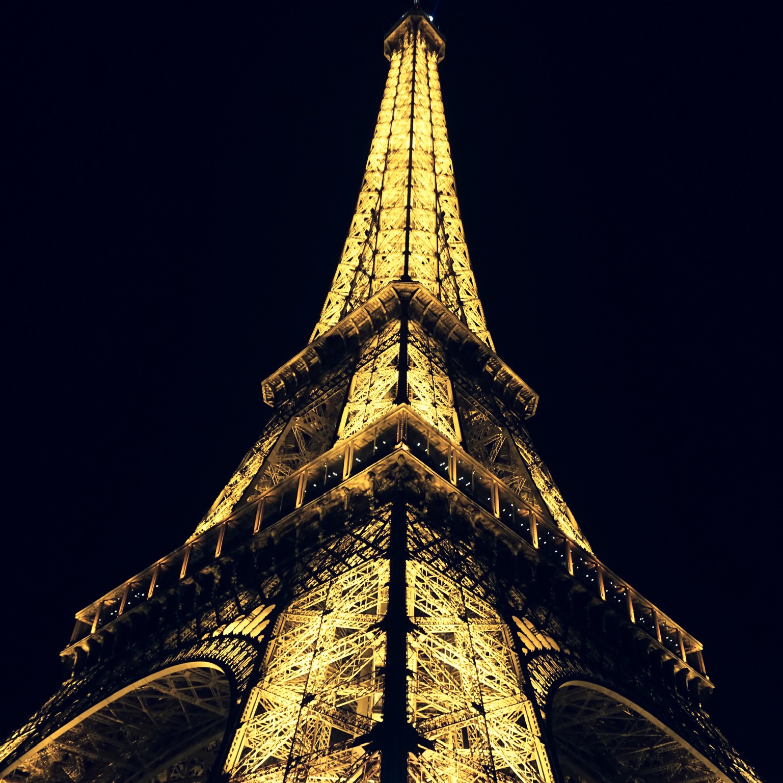 The Eiffel Tower blazing gold against a pure black sky, shot looking straight up its iron lattice.
