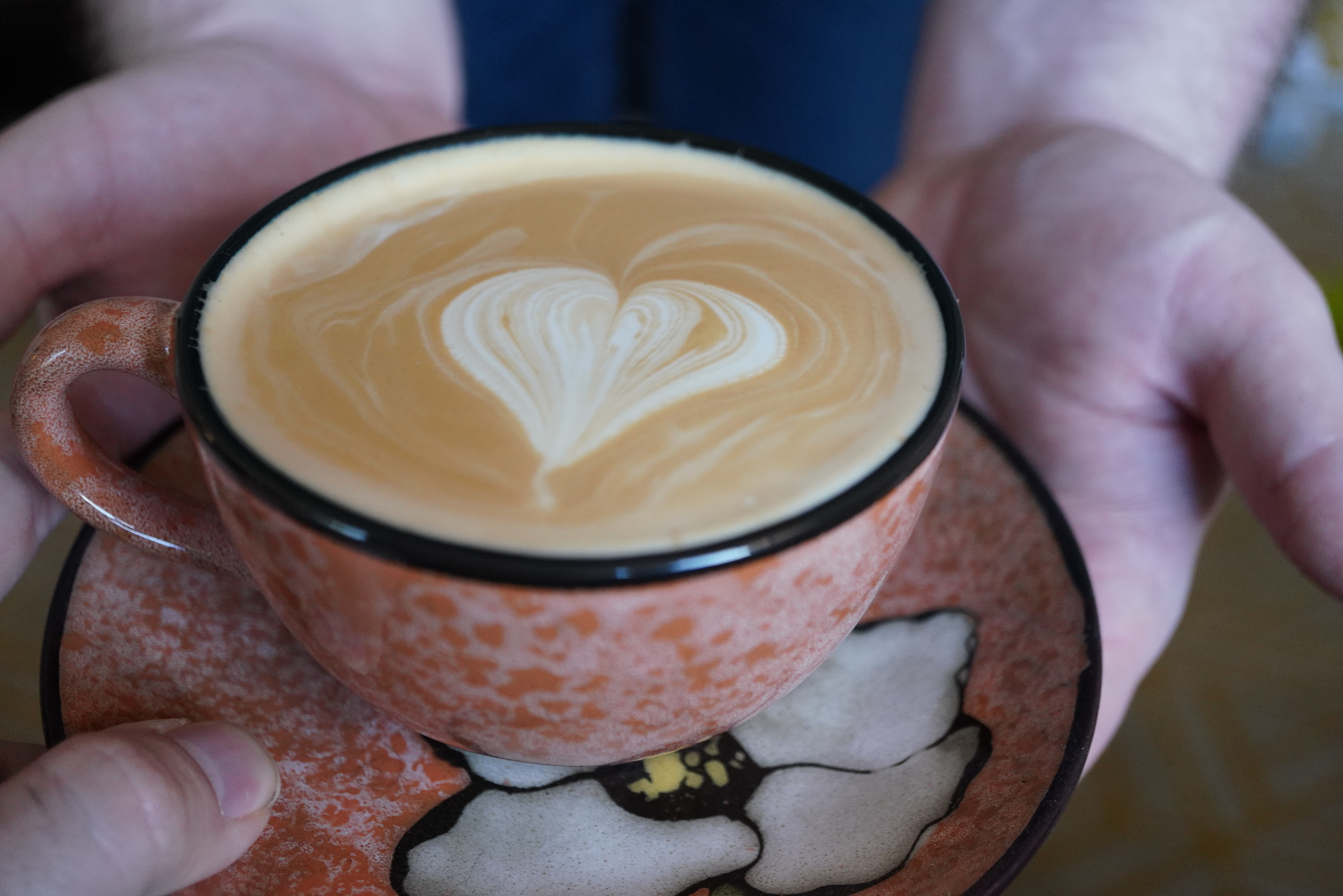 Two hands cradling an orange speckled ceramic cup with a heart poured in the foam.