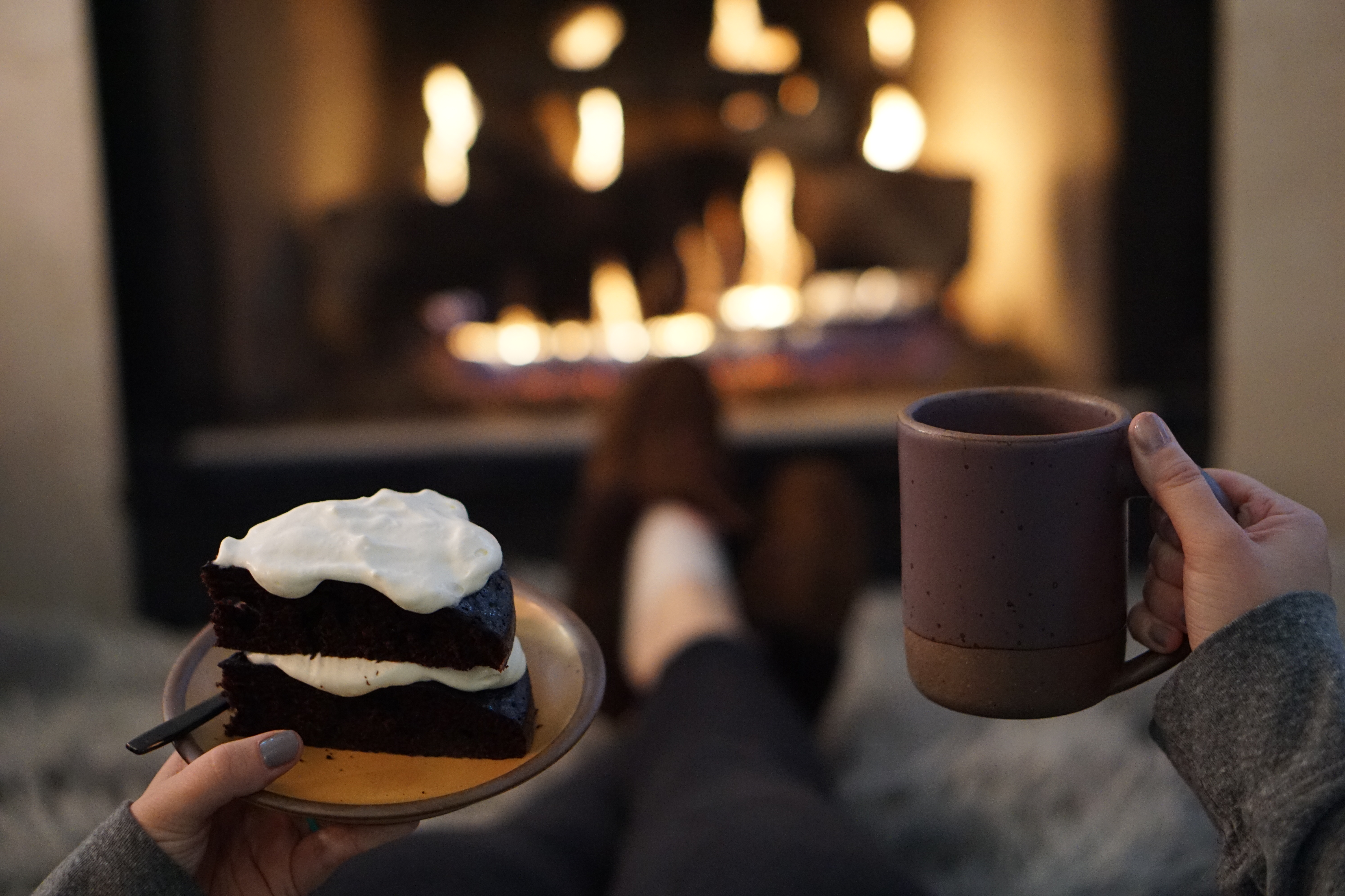 Hands holding a slice of dark chocolate cake and a speckled ceramic mug beside a glowing fireplace.