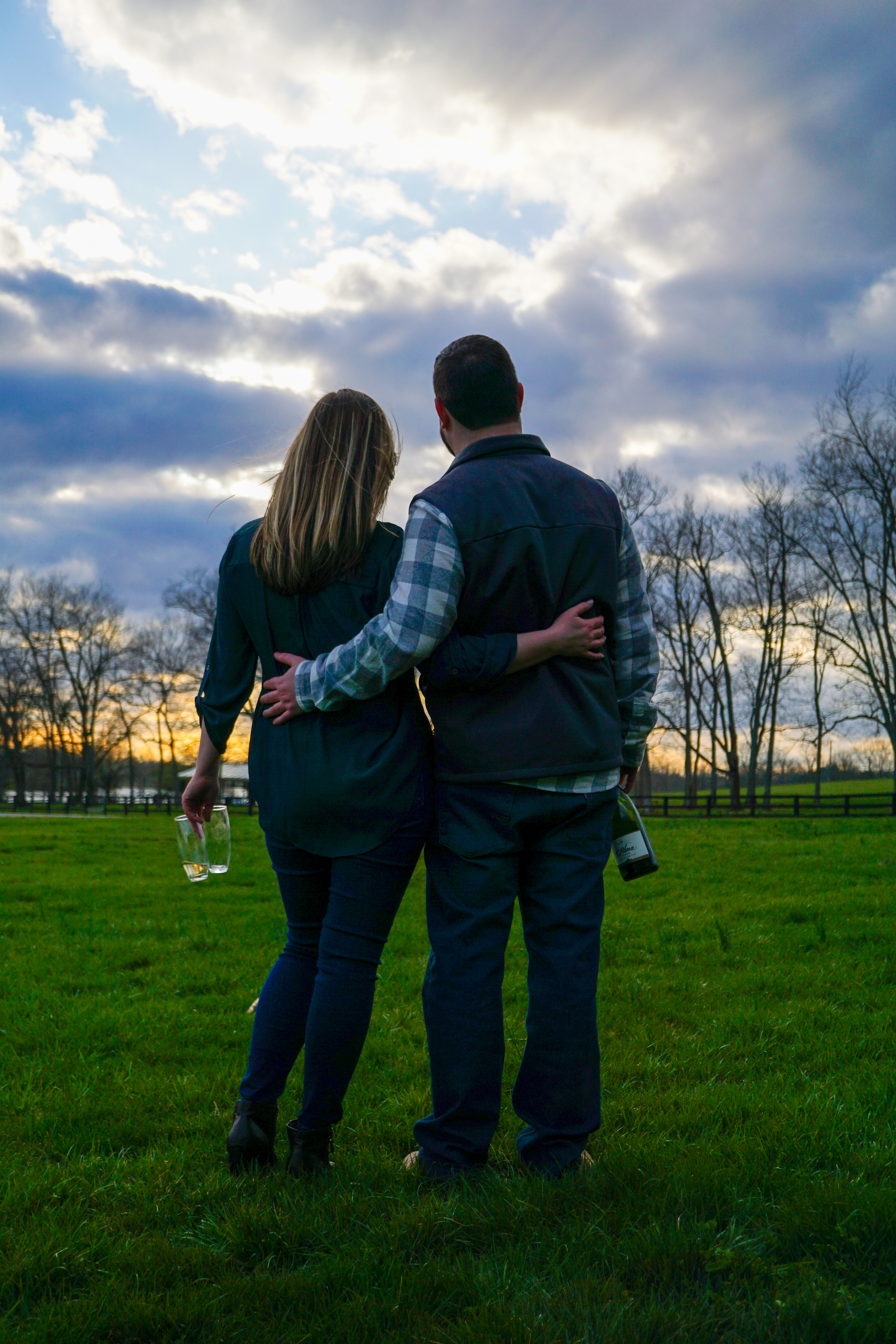 A couple standing arm in arm in a green field, facing a dramatic sunset sky with wine in hand.