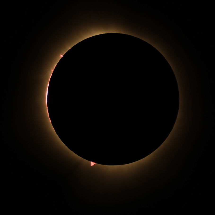Full totality with the corona glowing around the silhouetted moon, vivid red solar prominences erupting at the edge.