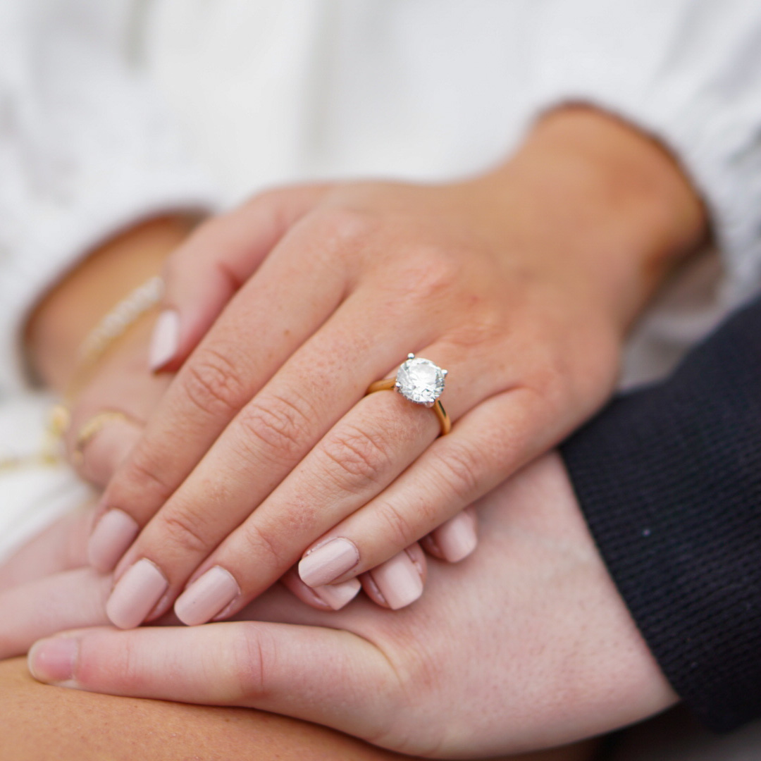 A close-up of intertwined hands, a round solitaire diamond on a yellow gold band catching soft light.